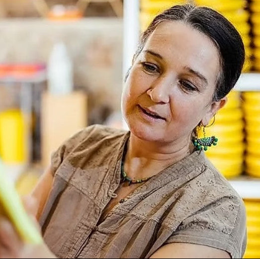 A close-up lifestyle shot of a woman with dark hair pulled back, wearing a brown textured short-sleeve top and green beaded earrings, looking down and smiling in what appears to be a workshop or studio.