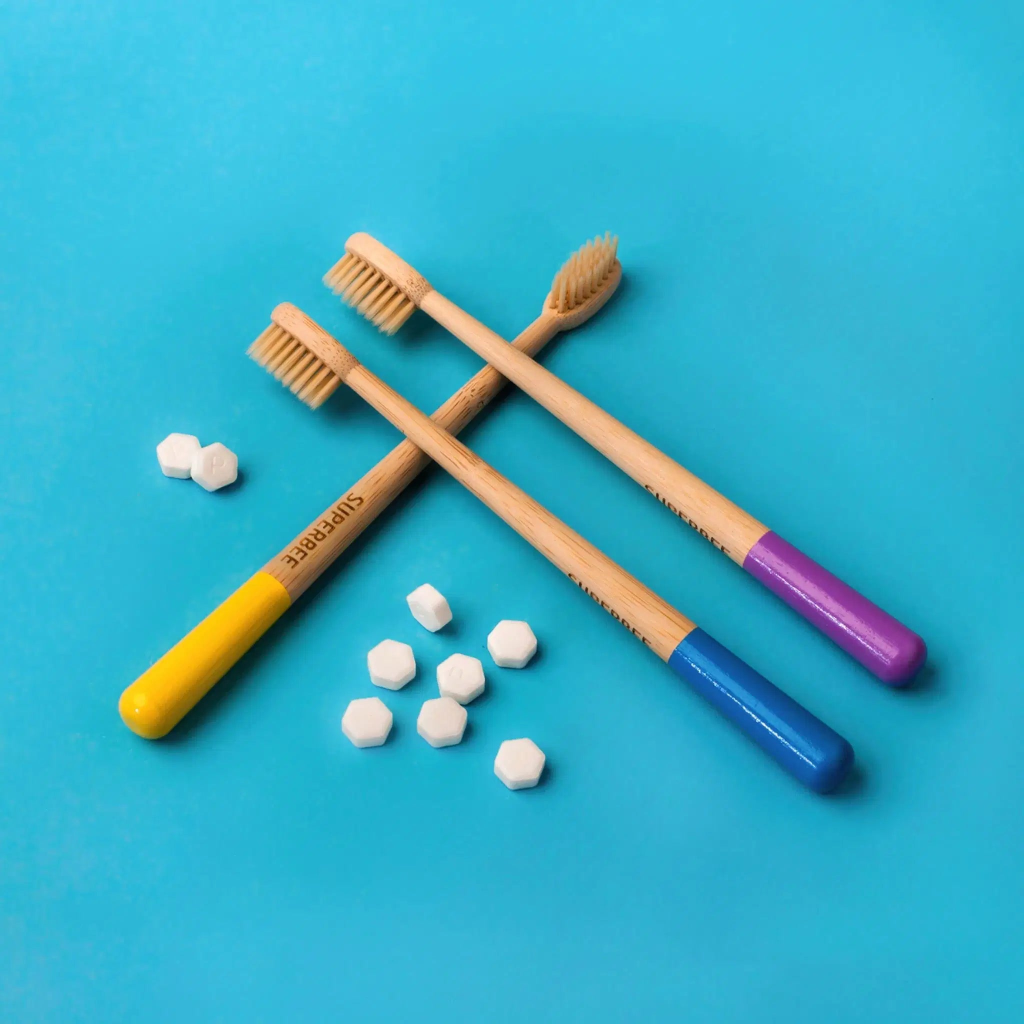 SuperBee Australia bamboo toothbrushes with yellow, blue, and purple handles and white toothpaste tablets on blue
