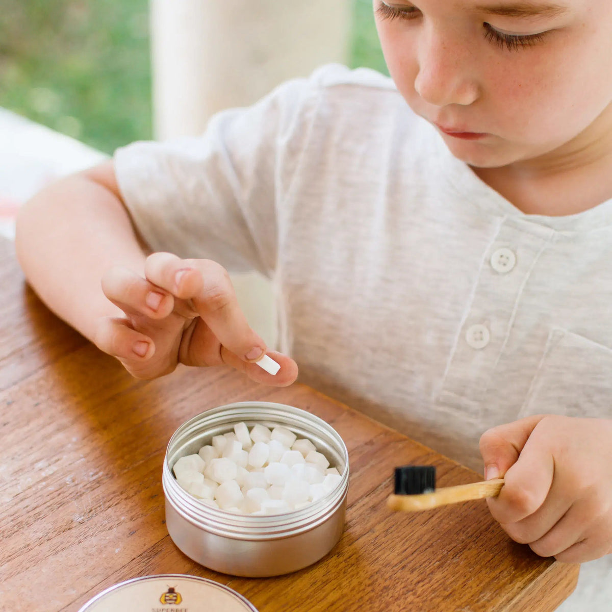 A young child holding a SuperBee Australia Dentos toothpaste tablet over a small metal tin with a bamboo toothbrush.
