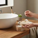 SuperBee Australia Dentos toothpaste tablets in a round cardboard container held by hands in a bathroom setting.