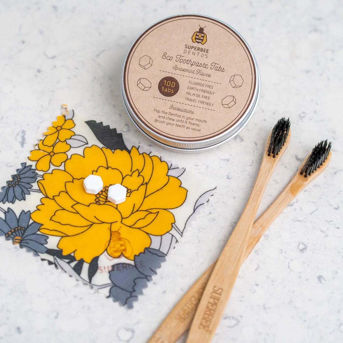 SuperBee Australia Dentos toothpaste tablets in a metal tin with two bamboo toothbrushes on a floral beeswax wrap.
